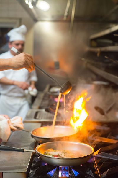 kitchen shot with frying pans over high flame, chef ladling sauce into one pan